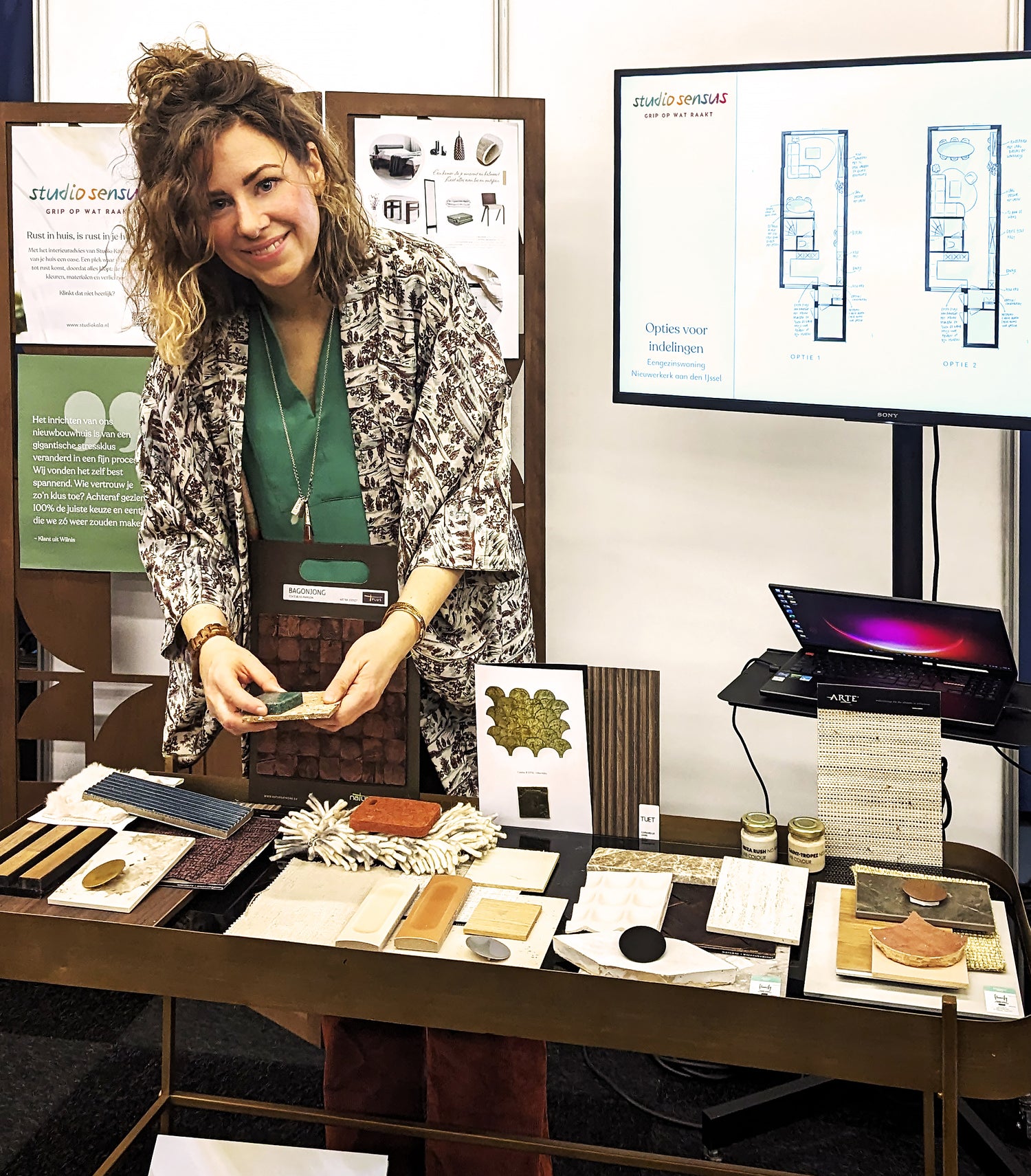 Michelle staat op een beurs met een stand vol met materiaalstalen en kijkt lachend op