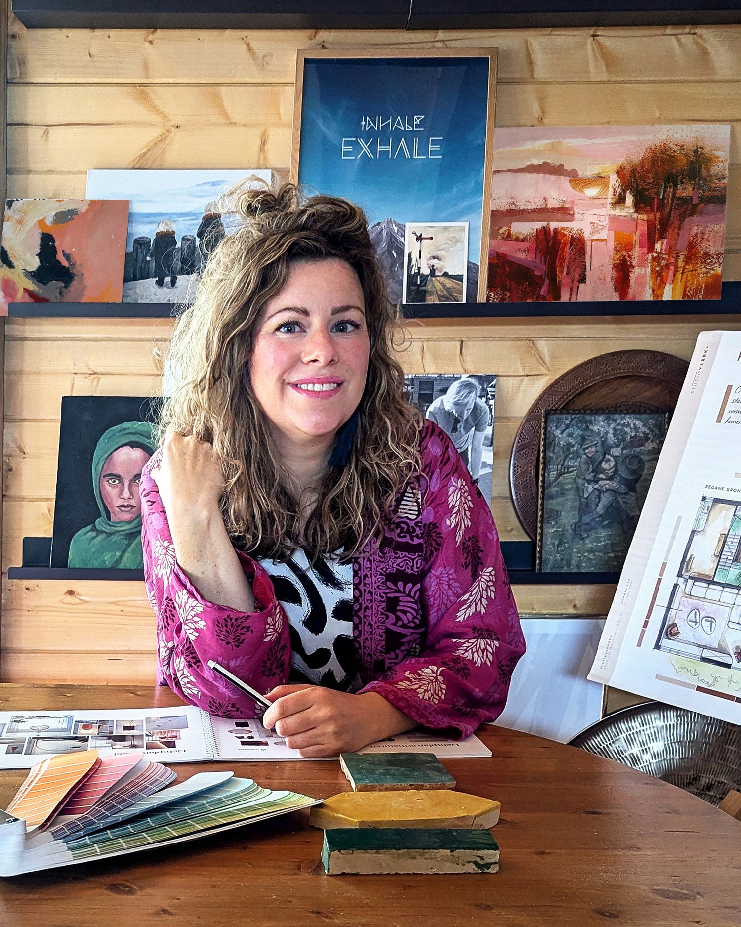 Michelle in haar studio, met voor zich een kleurenwaaier en tegeltjes. Achter haar staan kunstwerken op wandplanken.
